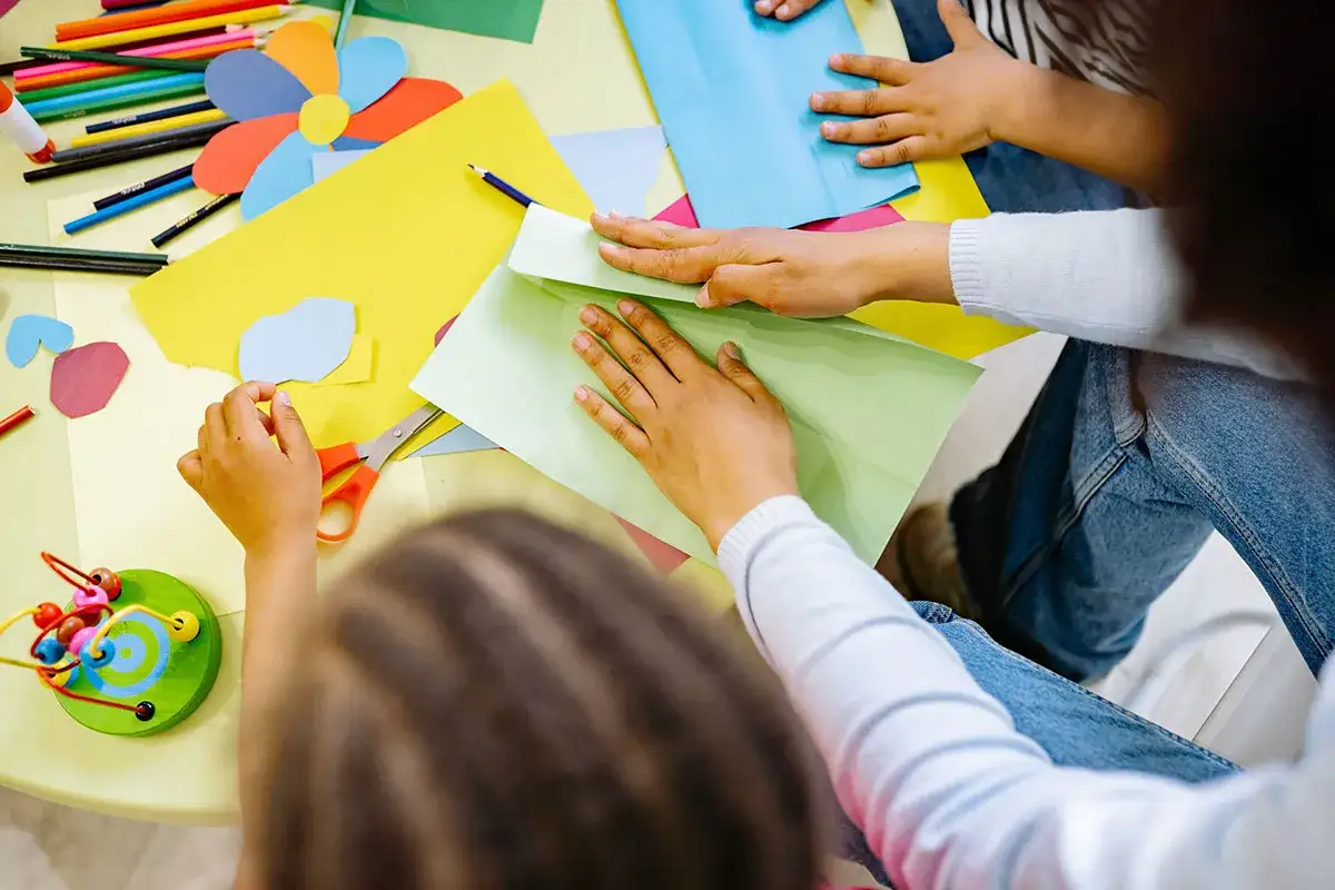 Des mains d'enfants pliantes du papier vert, entourées de crayons et de ciseaux, lors d'une activité créative au centre de loisirs 2 ans.