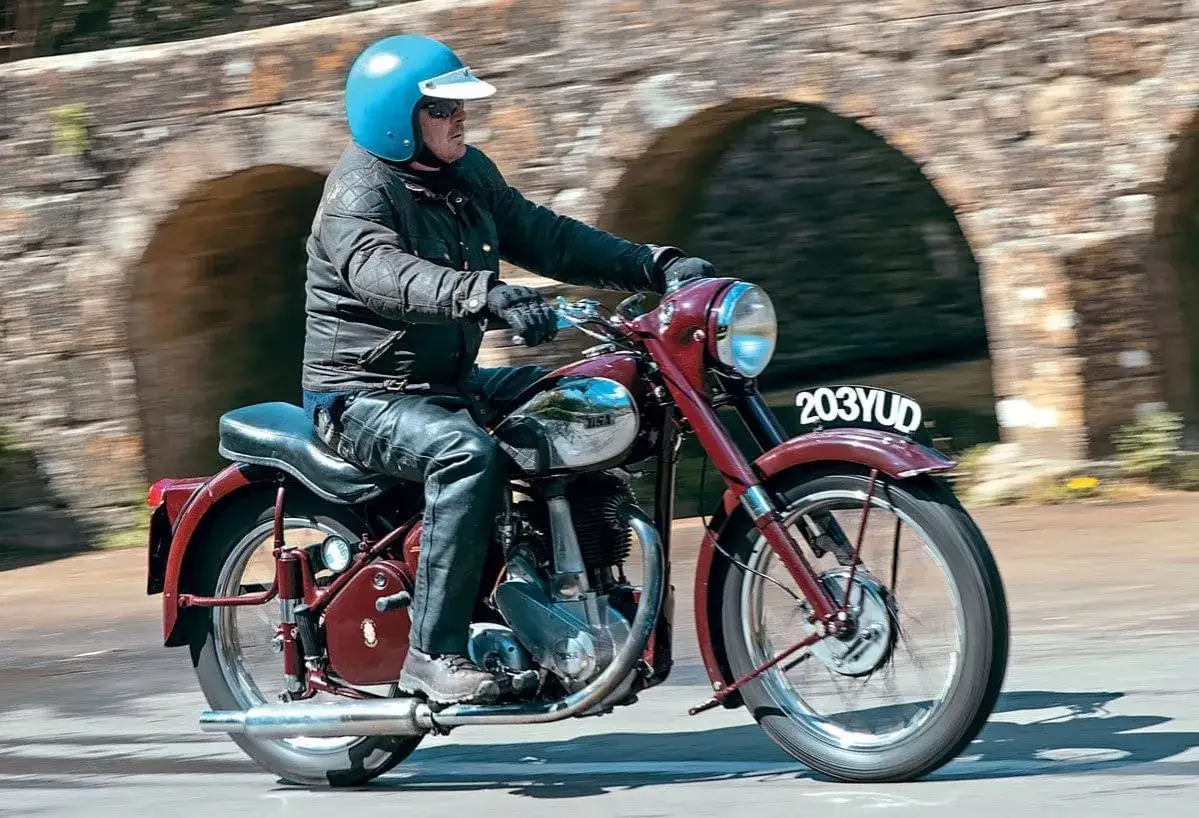 Mężczyzna w niebieskim kasku i sk&oacute;rzanej kurtce jedzie klasycznym, bordowym bsa motocykl pod kamiennym mostem.