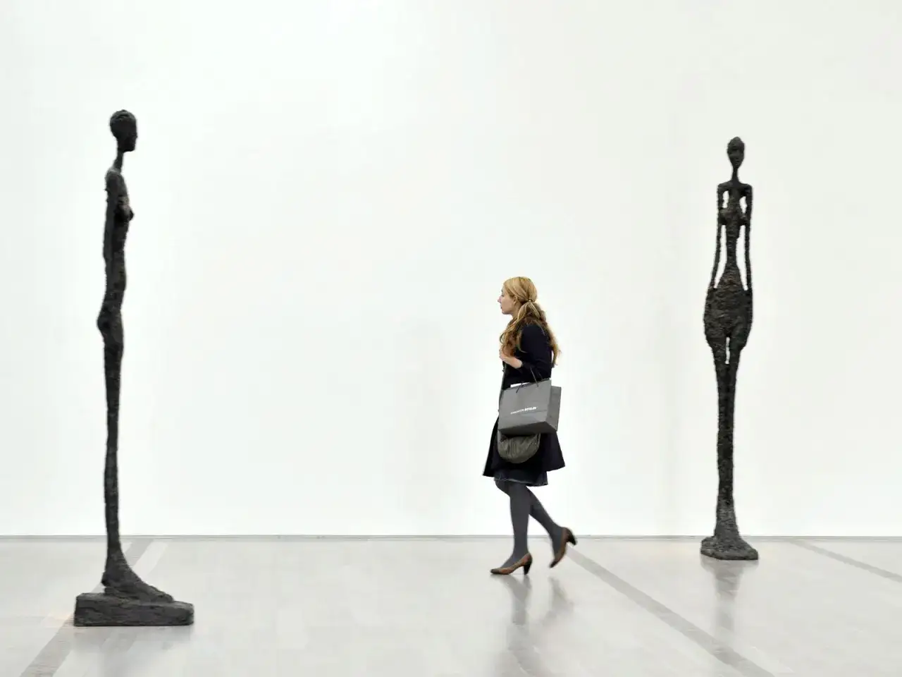 Une femme passe devant deux silhouettes filiformes, des sculptures embl&eacute;matiques de Giacometti, dans une galerie d'art &eacute;pur&eacute;e.