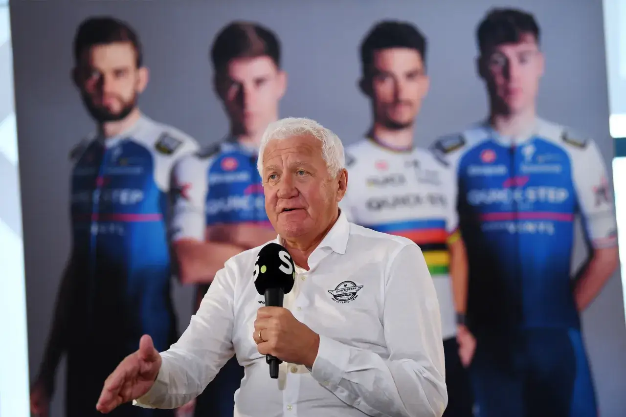 Patrick Lefevere, holding a microphone, speaks with four cyclists in Quick-Step Alpha Vinyl team jerseys blurred in the background.