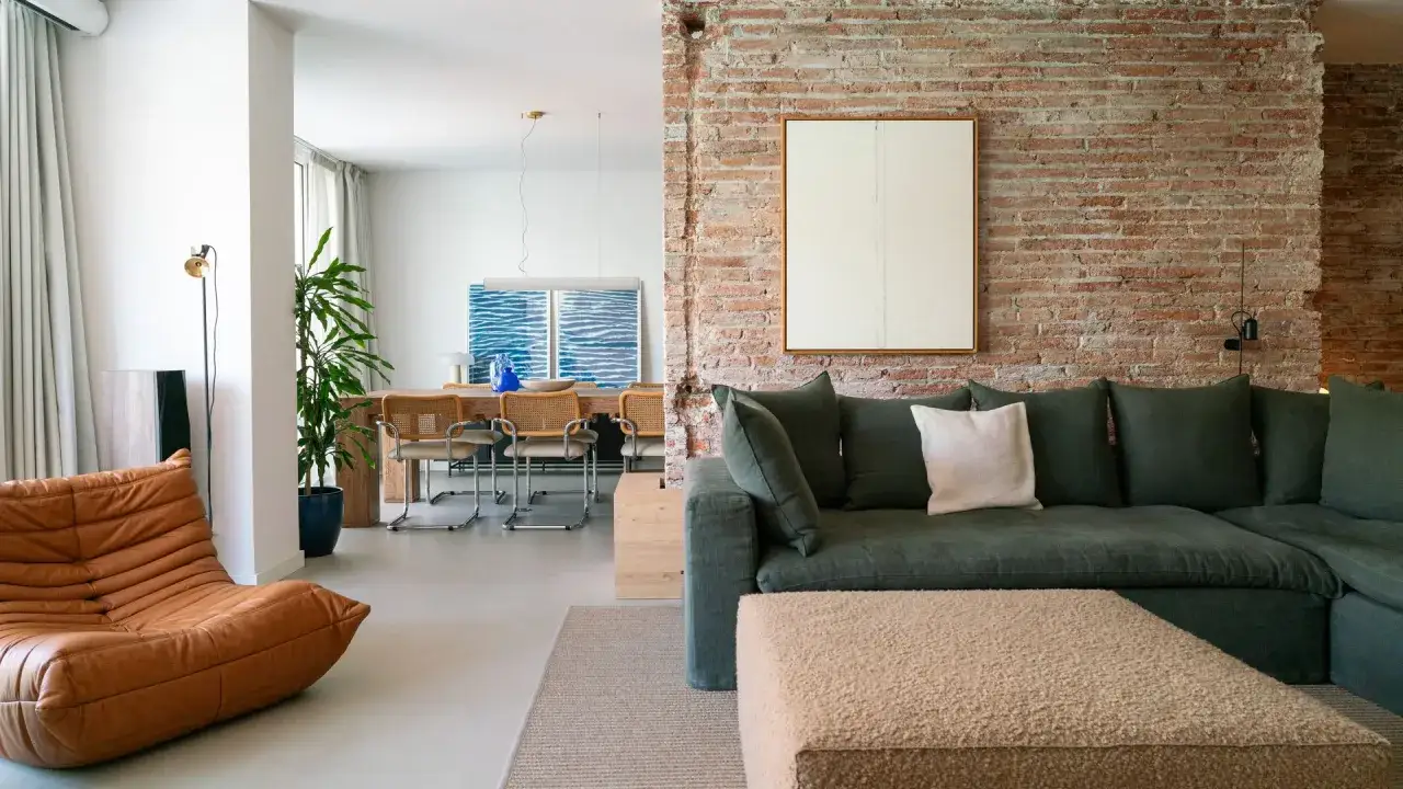 Sal&oacute;n moderno con sof&aacute; verde, sill&oacute;n de cuero y suelo de microcemento acabado. Pared de ladrillo visto y mesa de comedor al fondo.