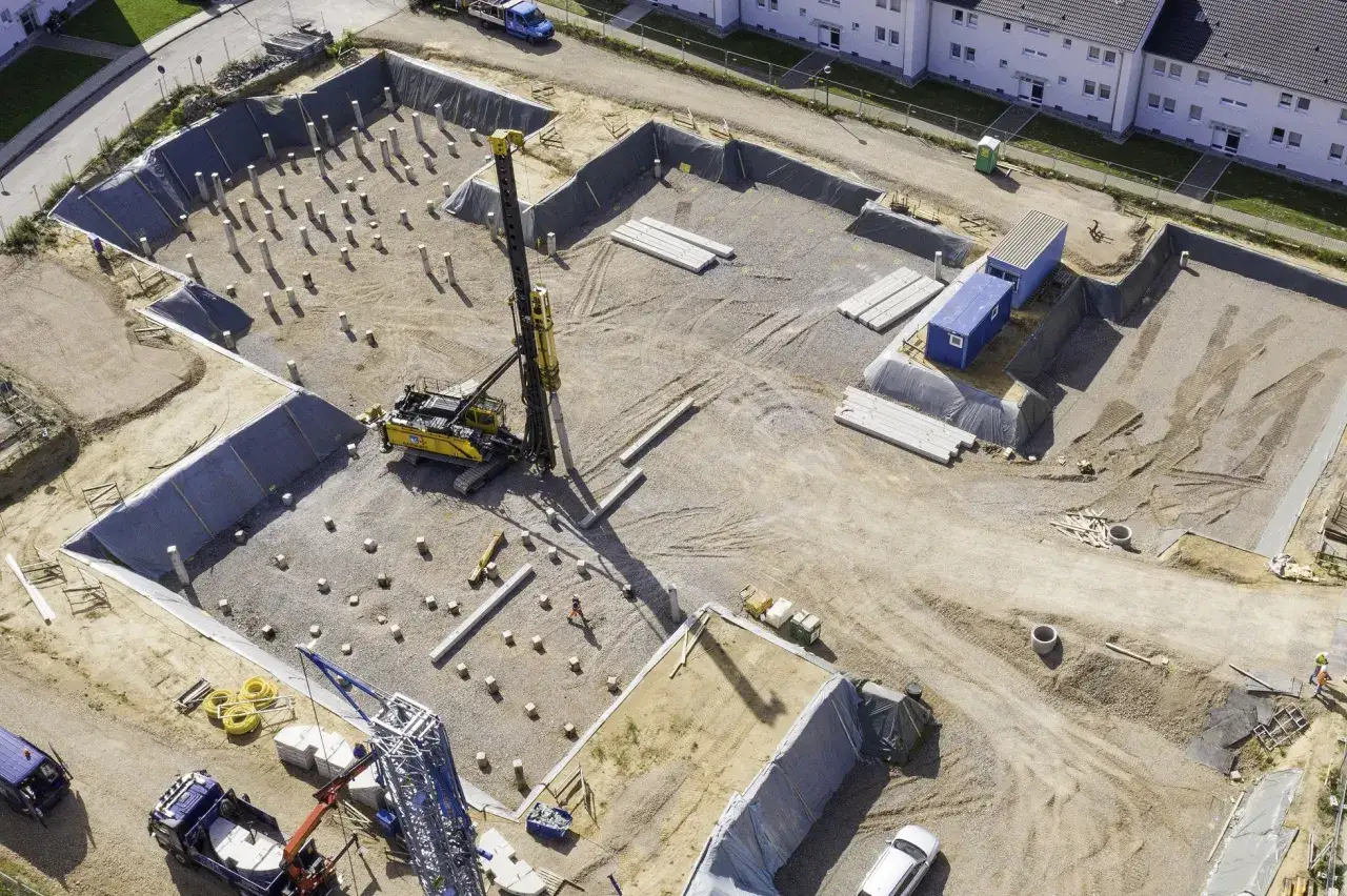 Plac budowy z koparką i rzędami betonowych pali. Powstaje tu dom na palach betonowych, fundamenty są przygotowywane.