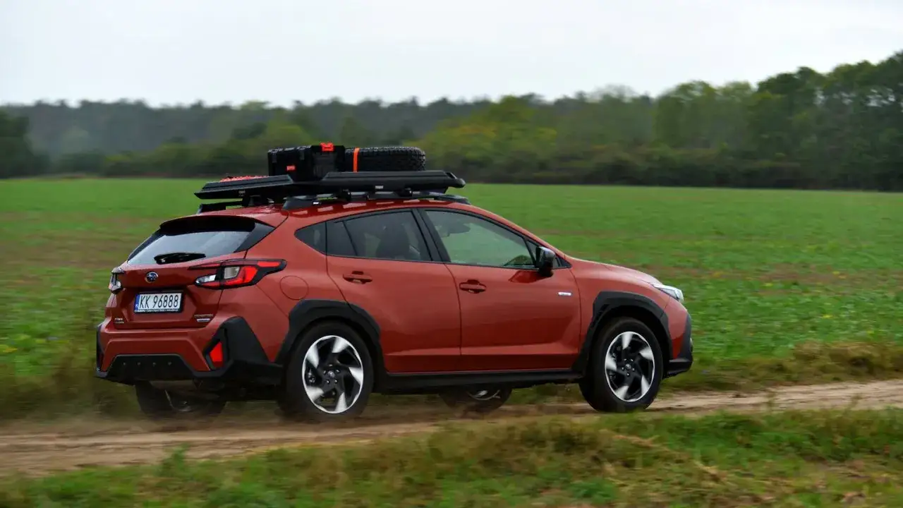Pomarańczowy Subaru Crosstrek 2025 z bagażnikiem dachowym i zapasowym kołem jedzie po polnej drodze wśród zielonych pól.