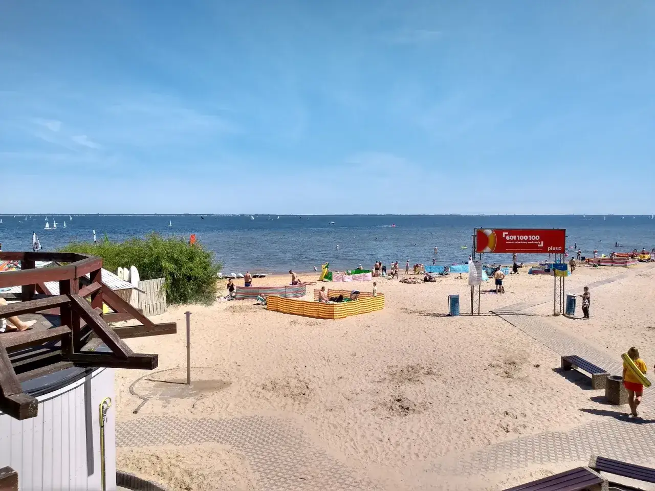 Słoneczna puck plaża z ludźmi odpoczywającymi na piasku i żagl&oacute;wkami na wodzie.