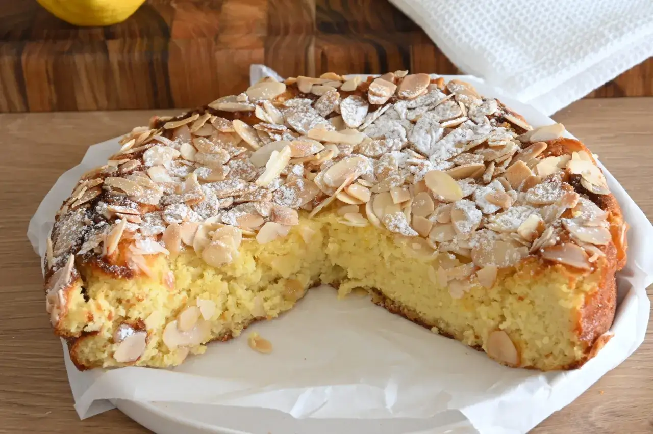 Torta delle monache dorata, cosparsa di mandorle a lamelle e zucchero a velo, con una fetta mancante che invita ad assaggiarla.
