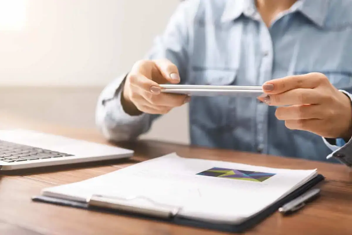 Persona usando su tel&eacute;fono para escanear documentos en una mesa con un port&aacute;til y un portapapeles.