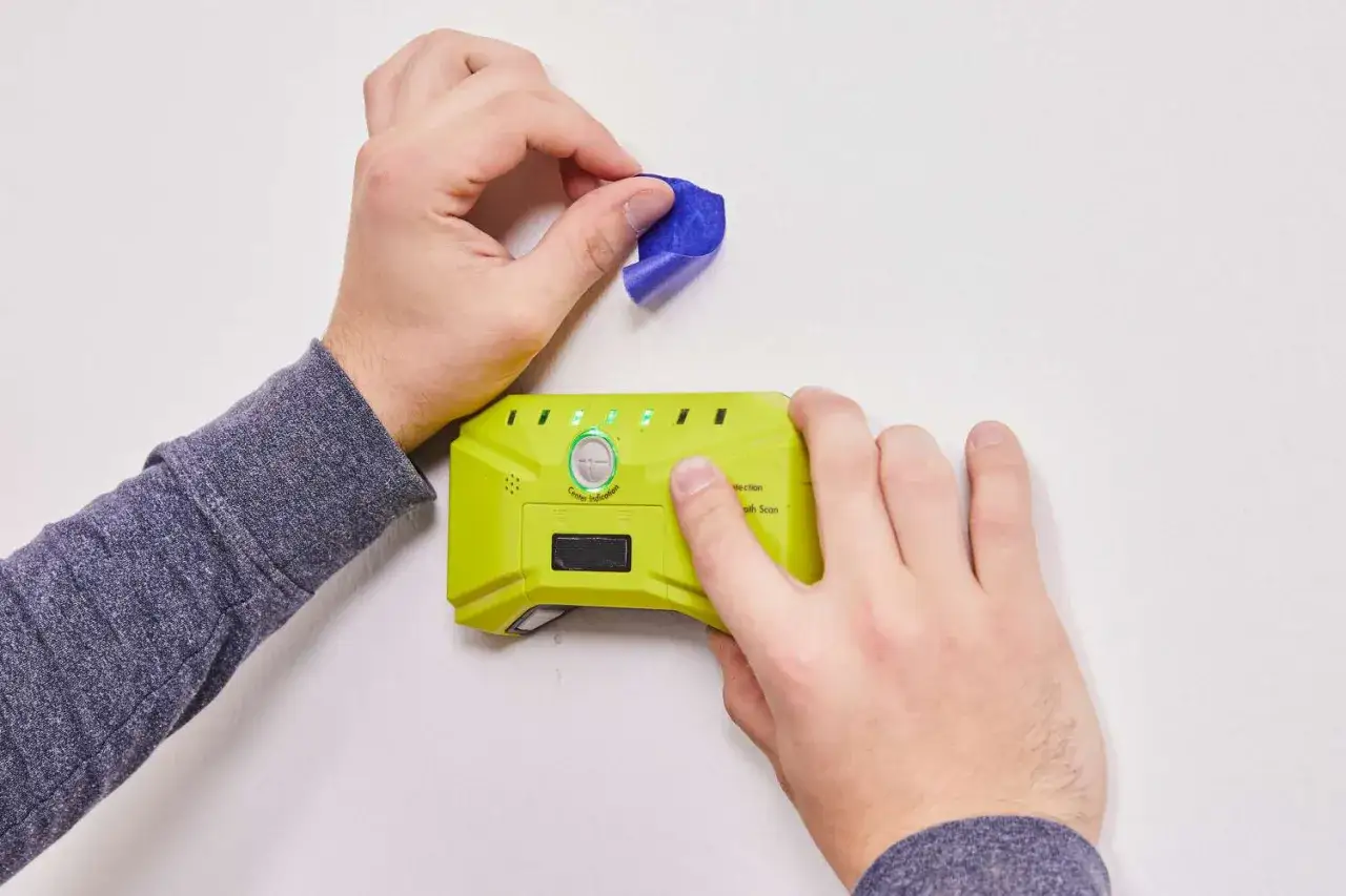 Using a stud finder is key to knowing how to hang shelves securely. A person uses blue tape to mark a spot on the wall while holding the device.