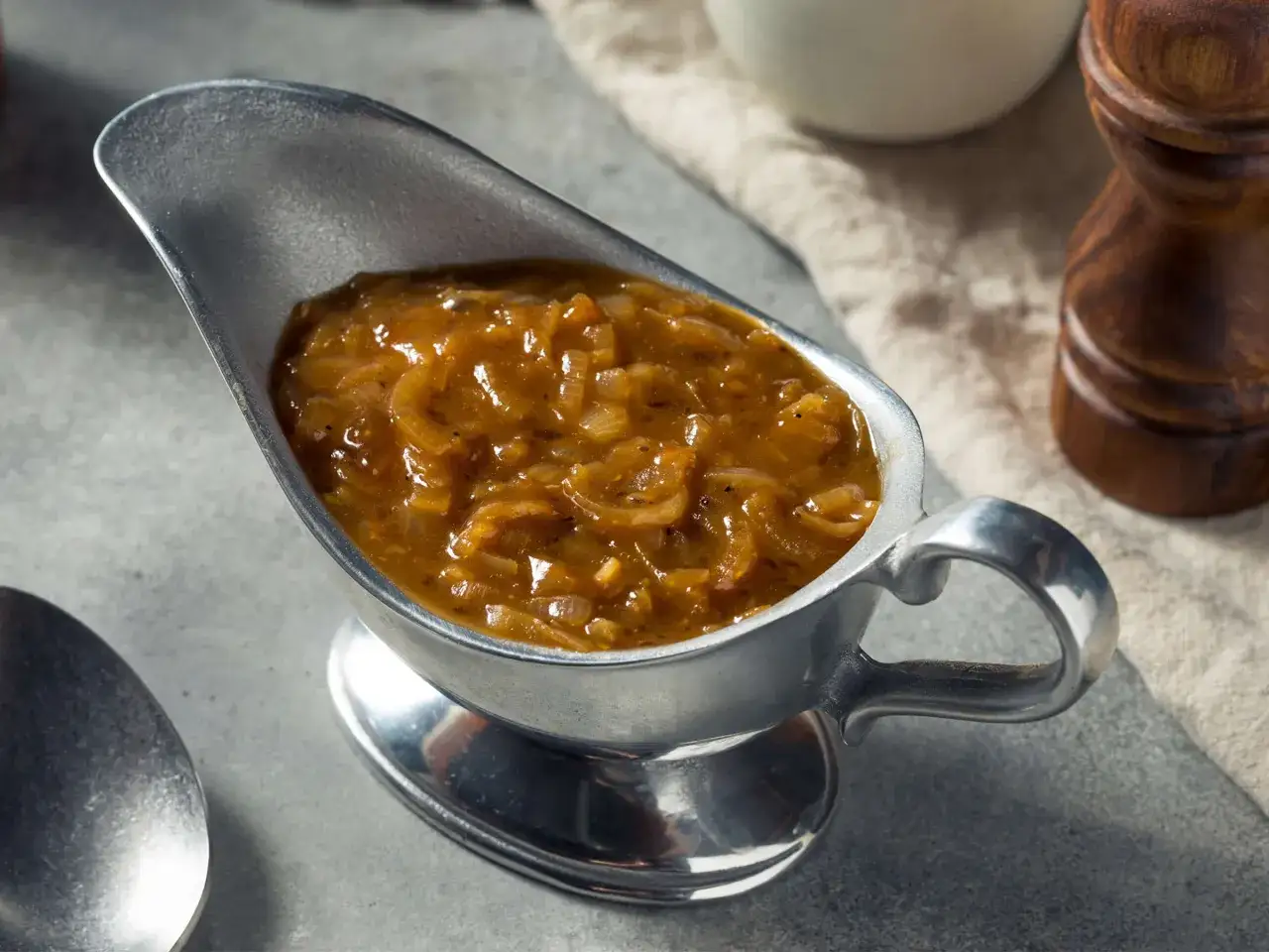Herzhafte zwiebelsauce wie bei Oma, serviert in einer silbernen Sauciere. Ein Löffel und ein Pfeffermühlen-Set deuten auf ein köstliches Essen hin.