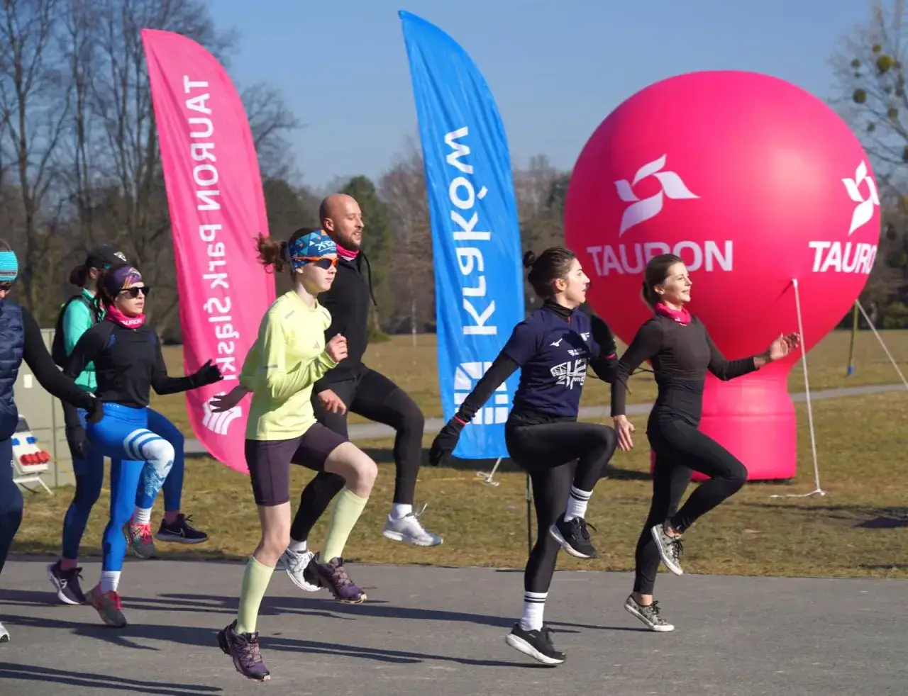 Grupa biegaczy ćwiczy, budując wydolność. W tle różowe i niebieskie flagi oraz duży, różowy balon z logo.