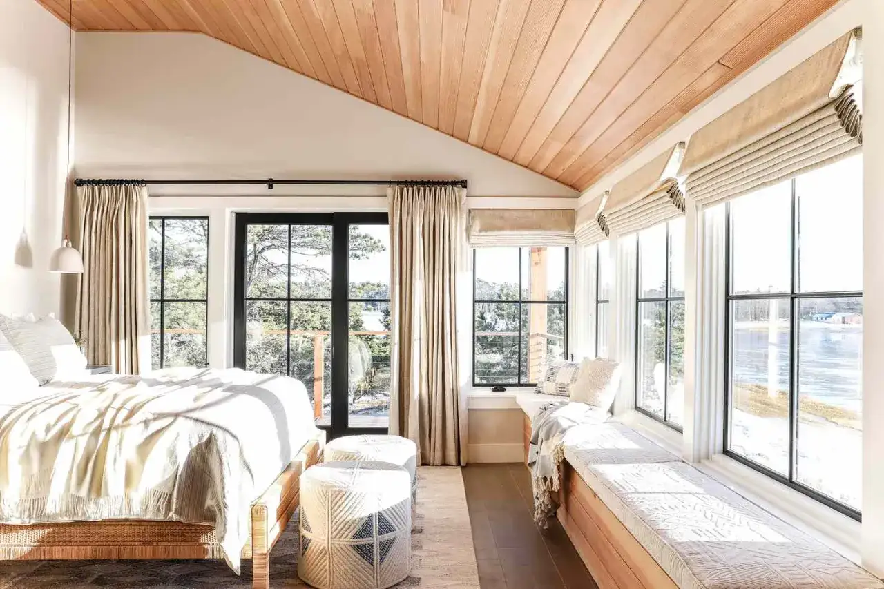 A cozy bedroom with a sloped, wood-paneled accent ceiling, large windows overlooking a wintery landscape, and a comfortable bed.