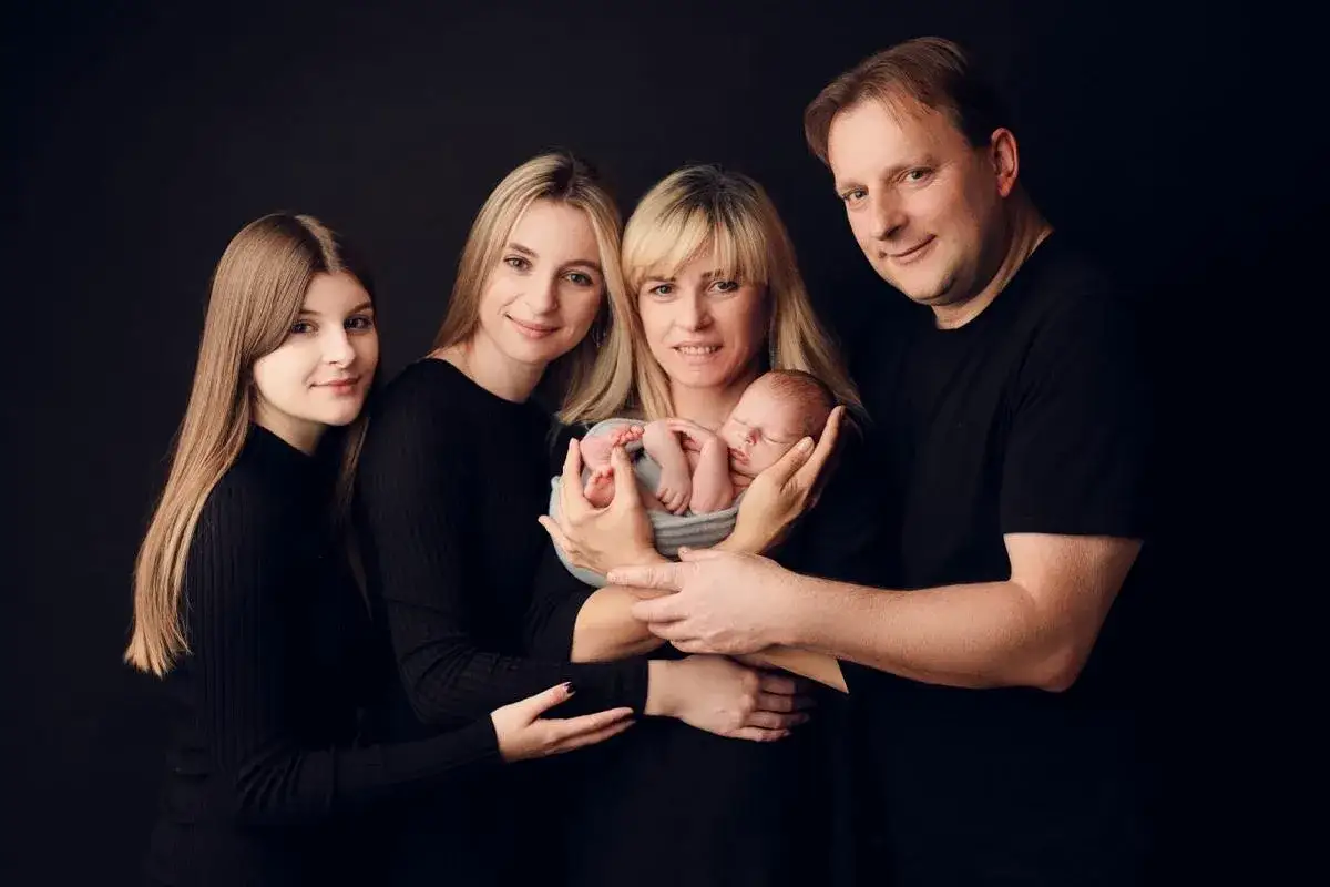 Rodzina z noworodkiem. Mama tuli dziecko, obok stoją starsza córka, młodsza córka i dumny ojciec. Wszyscy ubrani na czarno.