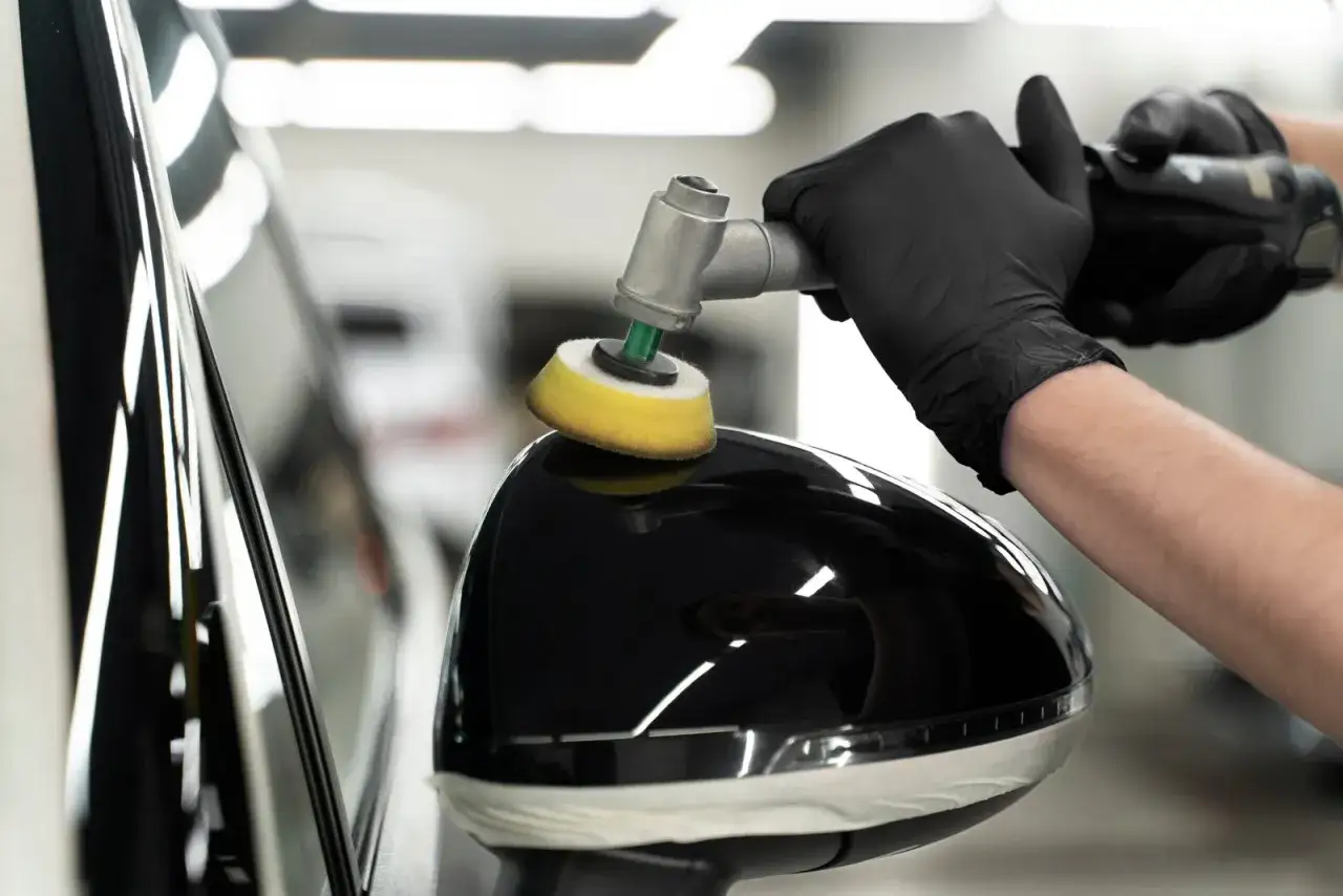 A gloved hand uses a polisher on a car's black side mirror. The process is to compound or polish first for a flawless finish.
