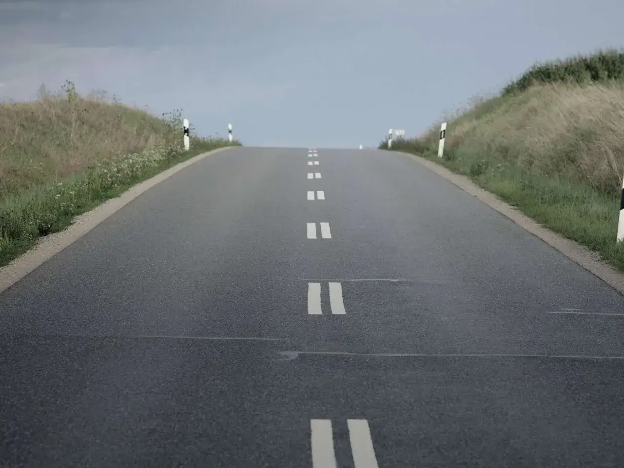 Eine Stra&szlig;e mit durchgezogener Linie f&uuml;hrt bergauf, ges&auml;umt von Gras und Leitpfosten.