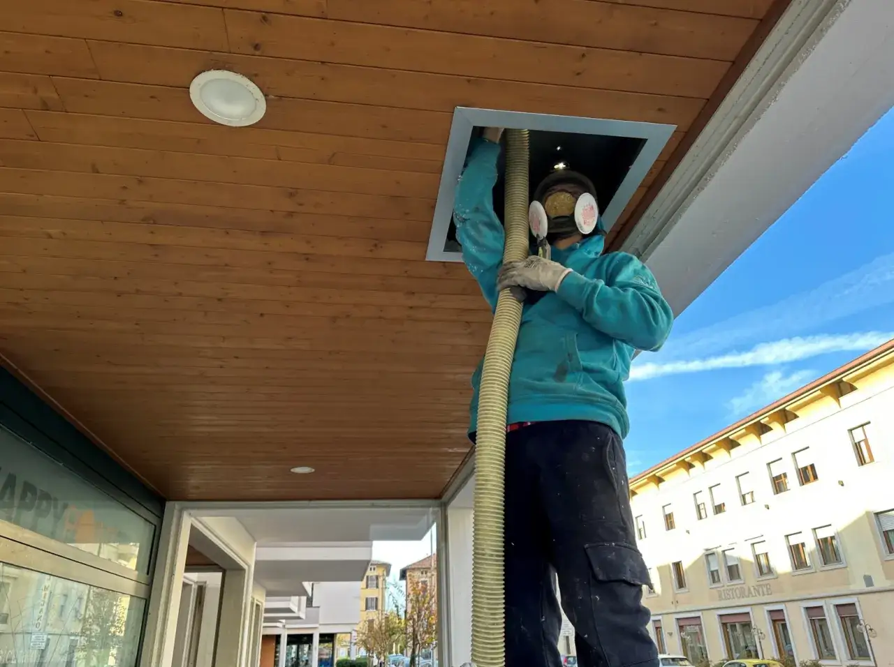 Operaio con maschera e guanti installa un controsoffitto isolante termico, insufflando materiale da un tubo.
