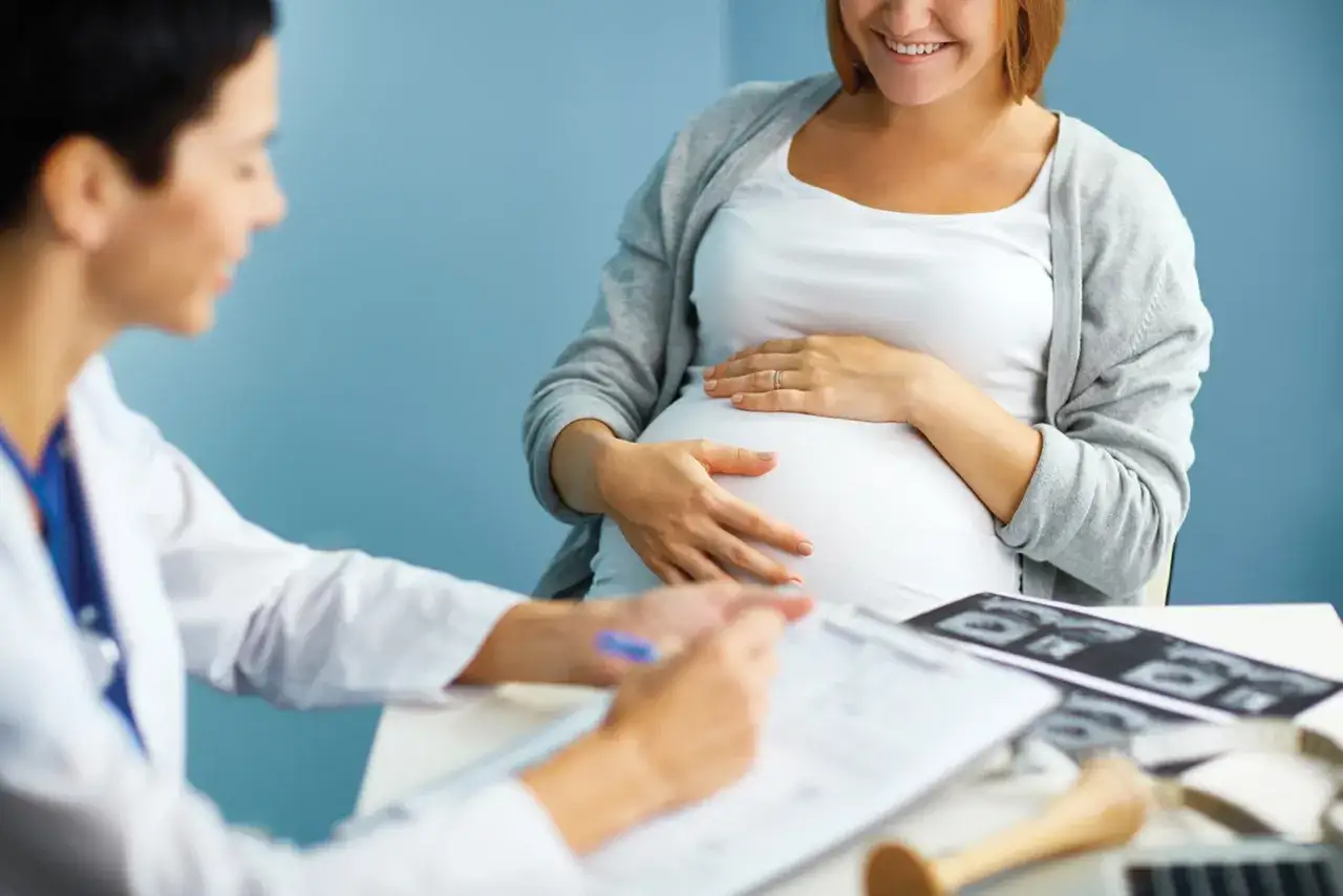 Une femme enceinte sourit en touchant son ventre, lors d'une consultation médicale. Le médecin écrit sur une tablette, un moment clé pour apprendre qu'on est enceinte.