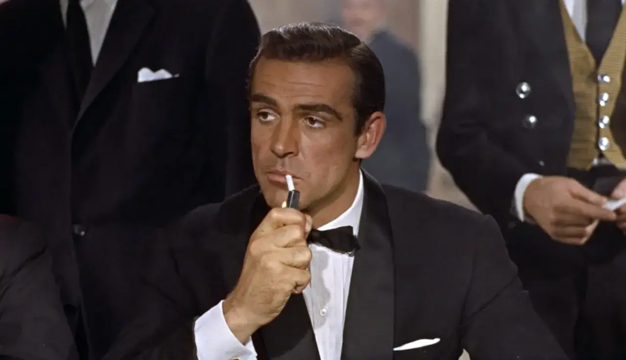 James Bond, elegante in smoking, accende una sigaretta con un'aria pensierosa.