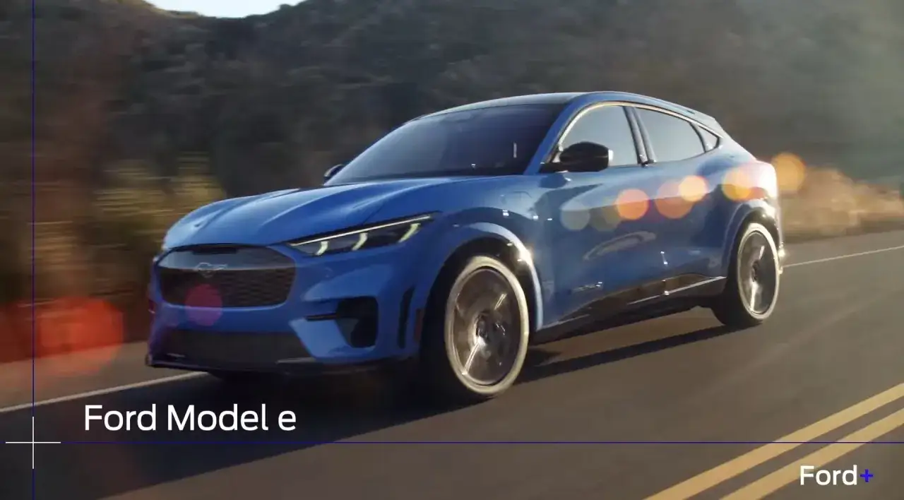 Niebieskie auto na e, Ford Mustang Mach-E, jedzie po krętej drodze wśród wzgórz.