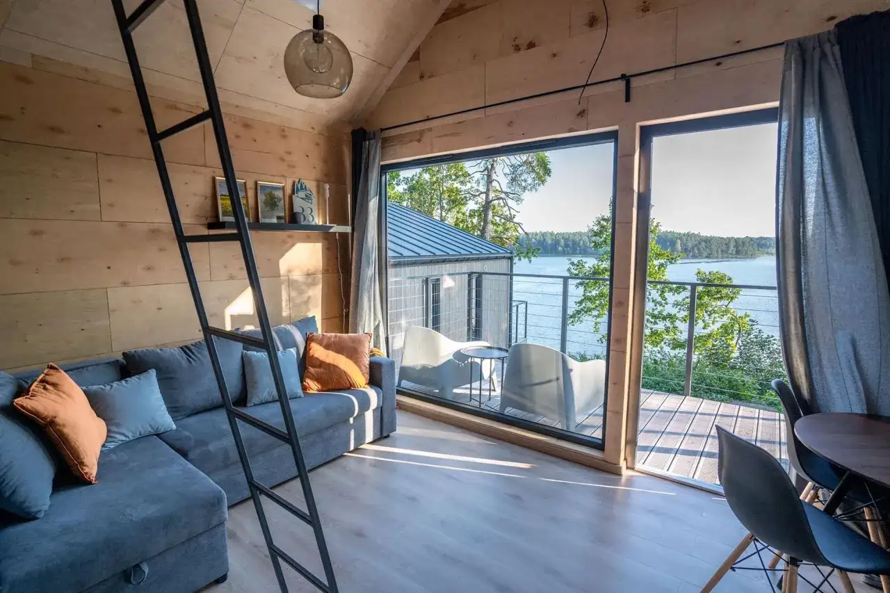Przytulne domki kempingowe Mazury z widokiem na jezioro. Sofa, drabinka na antresolę i taras z meblami.