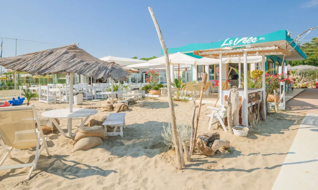 Un angolo di cervia mare con tavoli e sedie in spiaggia, ombrelloni di paglia e un chiosco colorato.