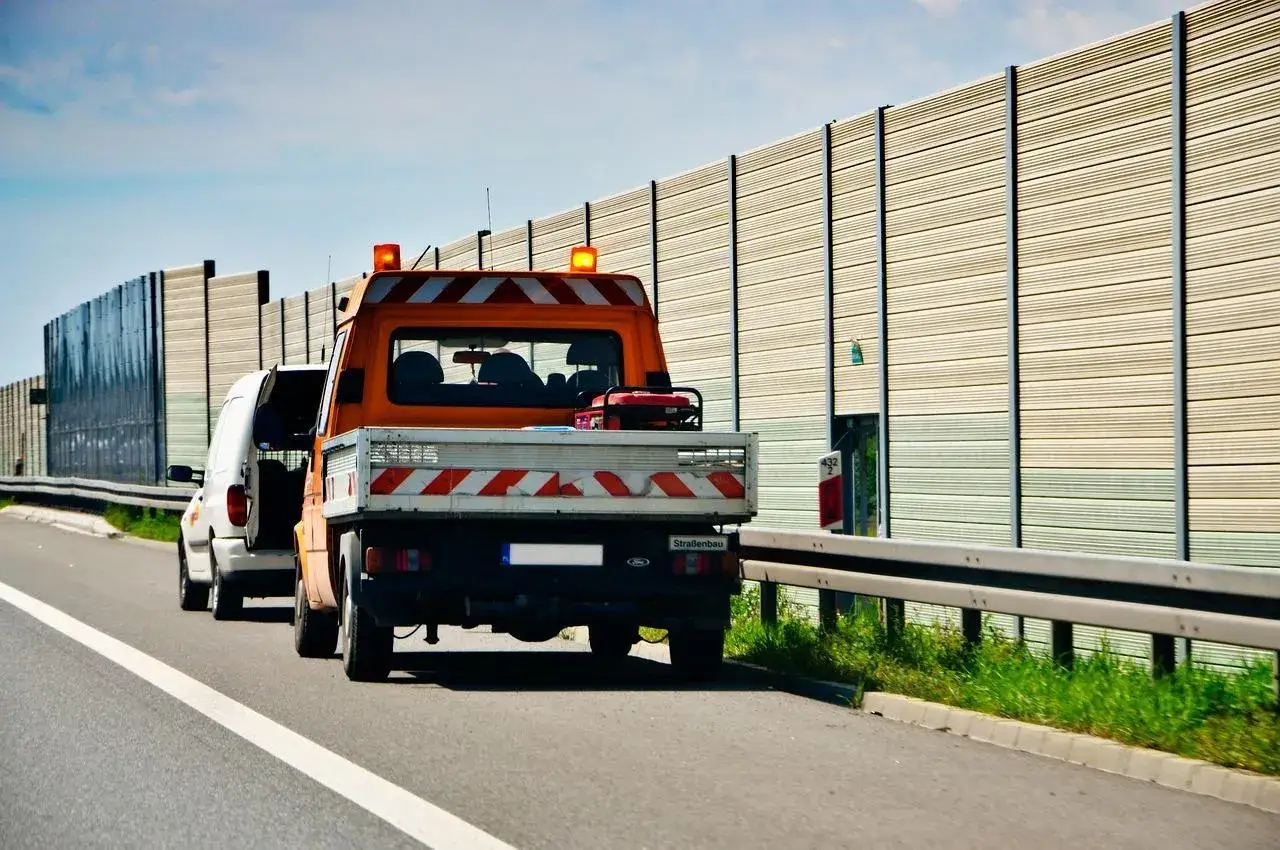 Un furgone bianco e un camion arancione con lampeggianti sono fermi sulla corsia d'emergenza. Ricorda che la distanza di sicurezza deve essere maggiore se si percorre la corsia d'emergenza.