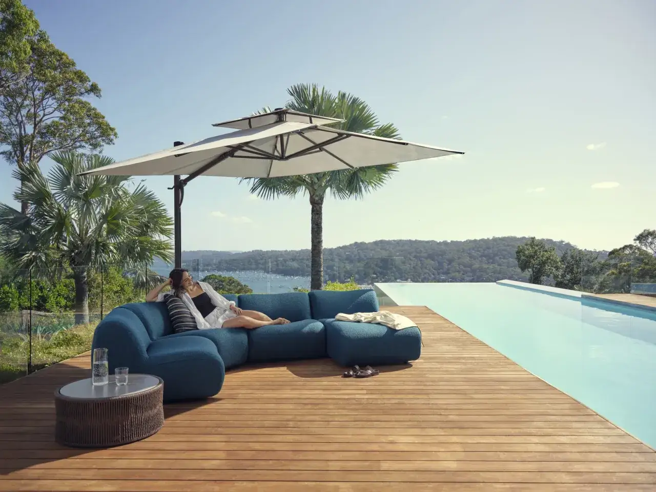 Femme détendue sur un canapé bleu, mobilier de jardin résistant aux intempéries, près d'une piscine à débordement avec vue sur la mer.