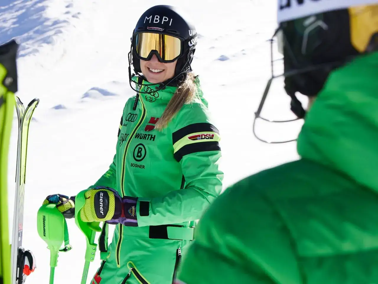 Narciarka w zielonym kombinezonie z logo Bogner, firmy znanej z odzieży sportowej, uśmiecha się na tle śniegu.