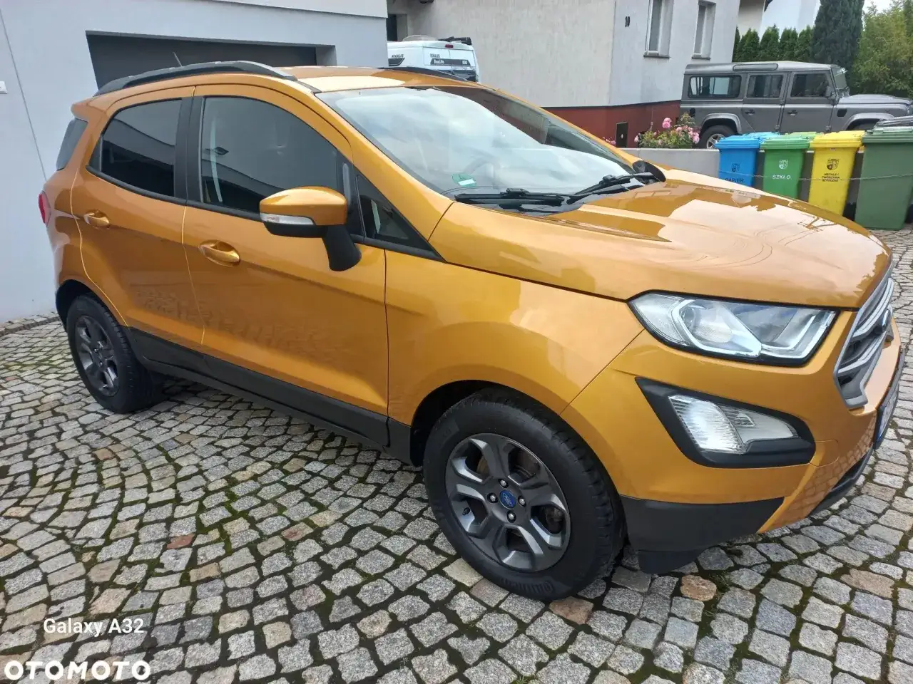 Złoty Ford Ecosport zaparkowany na brukowanym podjeździe. Obok stoją kolorowe kosze na śmieci.