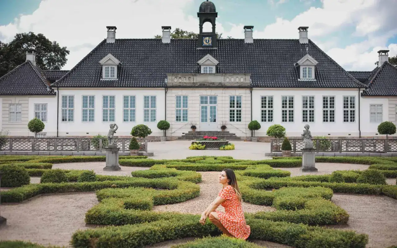 En kvinna i en orange klänning sitter i en vackert formad trädgård framför ett vitt slott i Danmark.