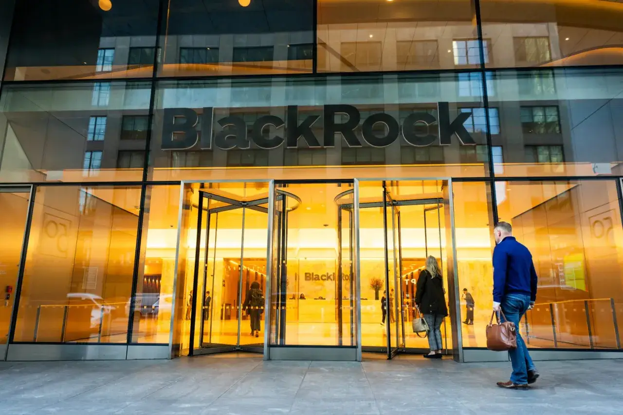 Wejście do siedziby BlackRock Polska. Szklana fasada budynku odbija światła i przechodni&oacute;w.