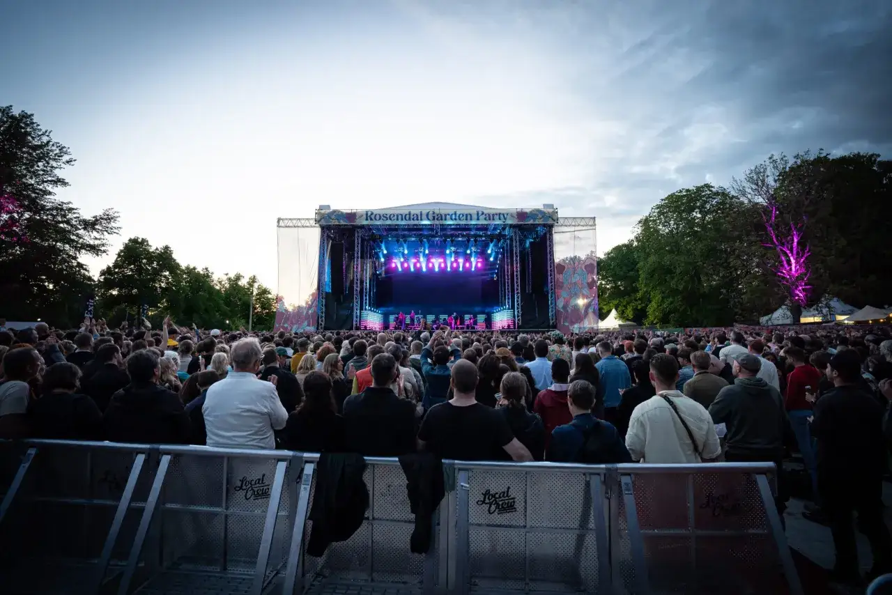 Publik njuter av en konsert vid Rosendal Garden Party. En stor scen med ljus och band syns i bakgrunden, omgiven av träd och en folkmassa.