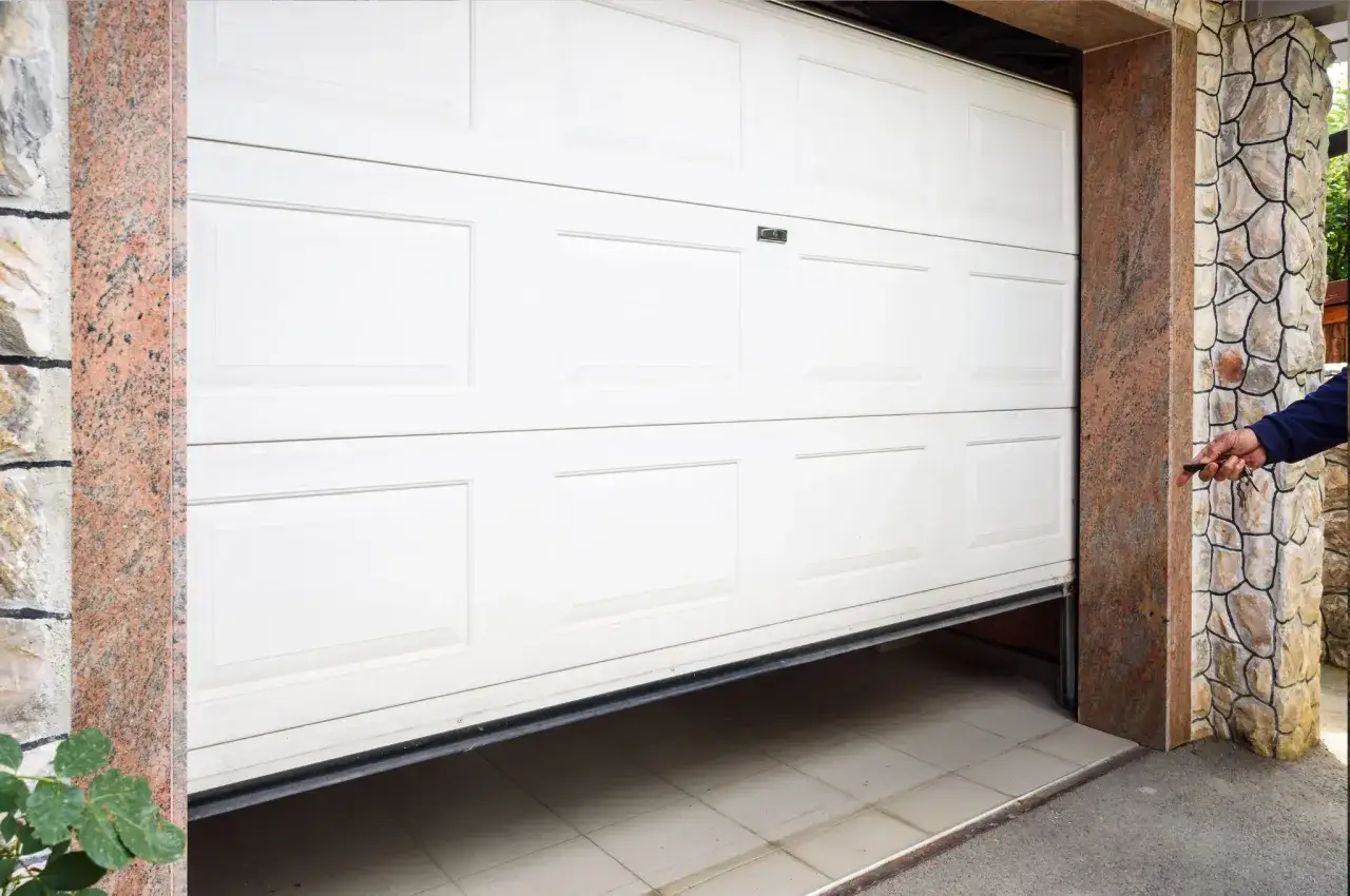 En person h&aring;ller i en fj&auml;rrkontroll f&ouml;r att byta garageport. Den vita porten &auml;r delvis &ouml;ppen.