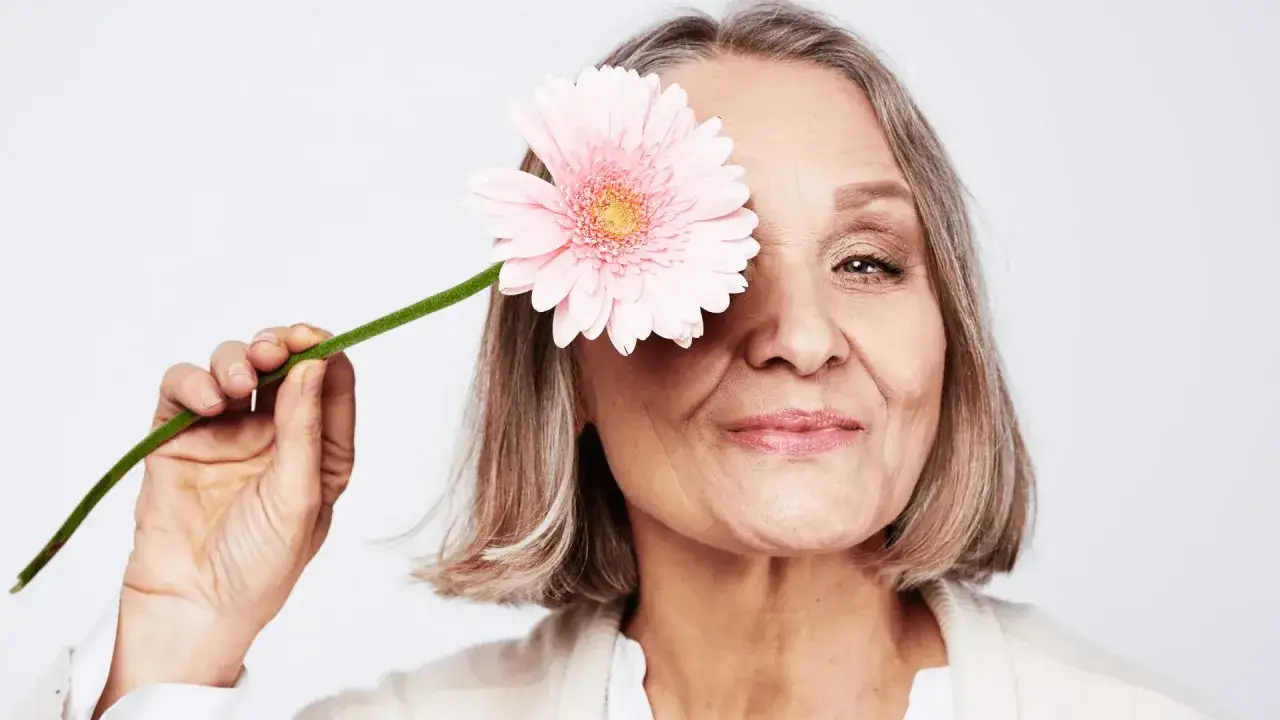 Uśmiechnięta dojrzała kobieta z różową gerberą zasłaniającą oko. Odkryj naturalne sposoby na menopauzę.