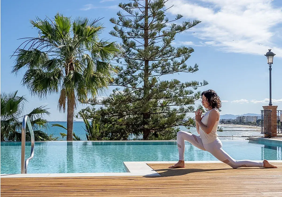 Mujer practica yoga junto a una piscina, un secreto para mantenerse joven. Palmeras y mar de fondo.
