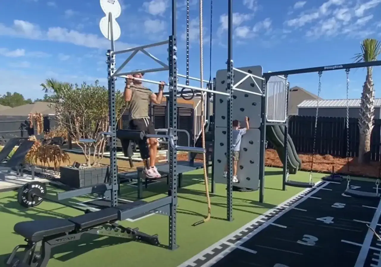 Un hombre hace dominadas en un gimnasio en el jardín. Un niño escala una pared. Hay equipo de entrenamiento y una pista de carreras.