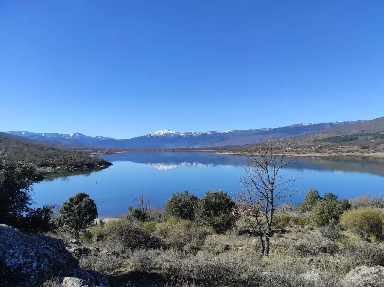 Paisaje sereno en el puerto de Canencia. Un lago azul refleja las monta&ntilde;as nevadas bajo un cielo despejado.