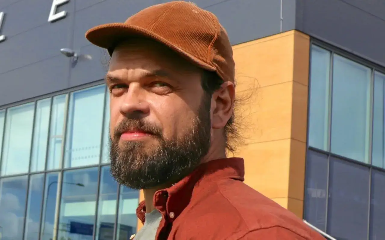 Marcus Berggren ung, med brun keps och röd skjorta, står framför en modern byggnad med stora fönster.
