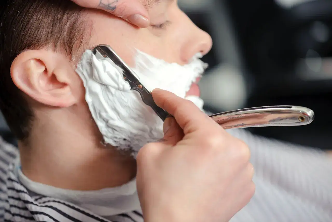 Barbero afeitando a contrapelo con navaja clásica, espuma blanca cubriendo la mejilla.