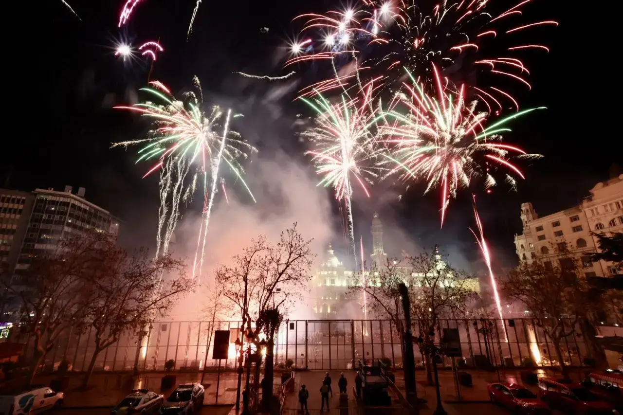 Espectacular fiesta en Valencia con fuegos artificiales iluminando el cielo nocturno sobre edificios hist&oacute;ricos y &aacute;rboles desnudos.