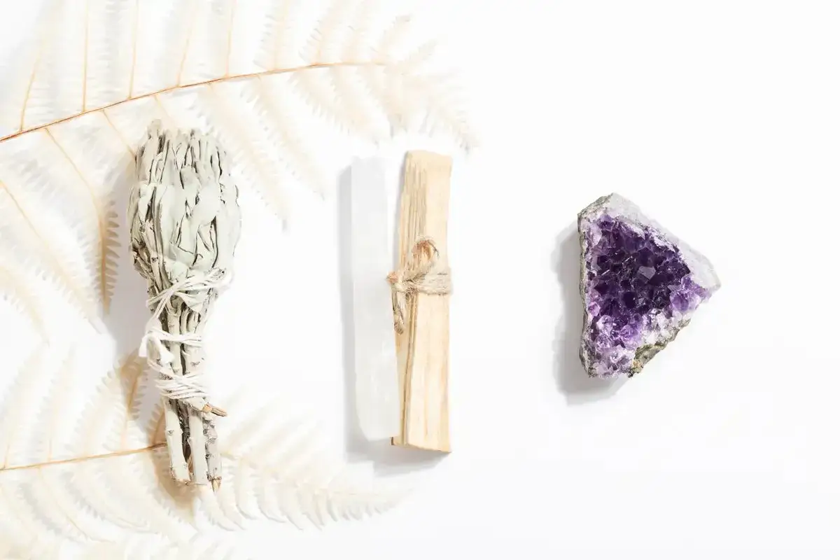 Un manojo de salvia, un palo santo atado con cuerda y una amatista sobre fondo blanco. Se puede quemar palo santo de noche para purificar el ambiente.