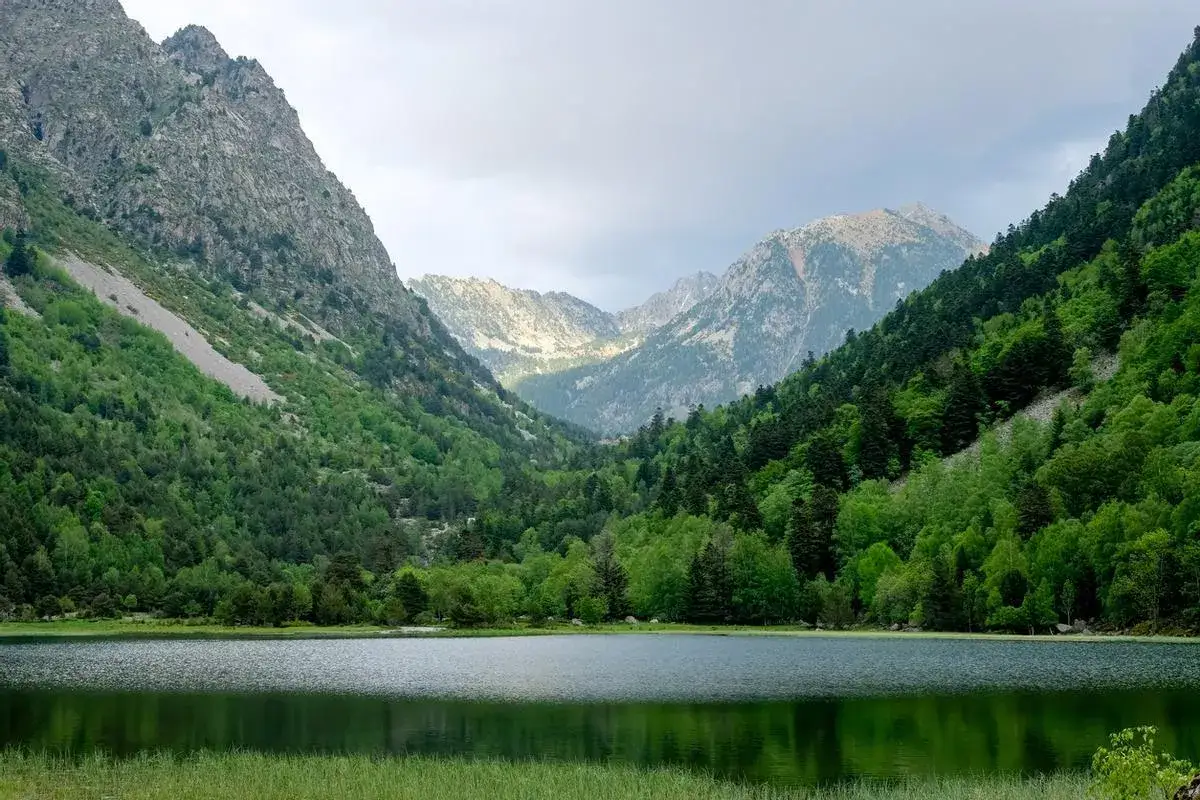 Paisaje sereno de los Lagos de Colomers, ideal para una ruta corta. Monta&ntilde;as verdes y un lago tranquilo reflejan el cielo nublado.