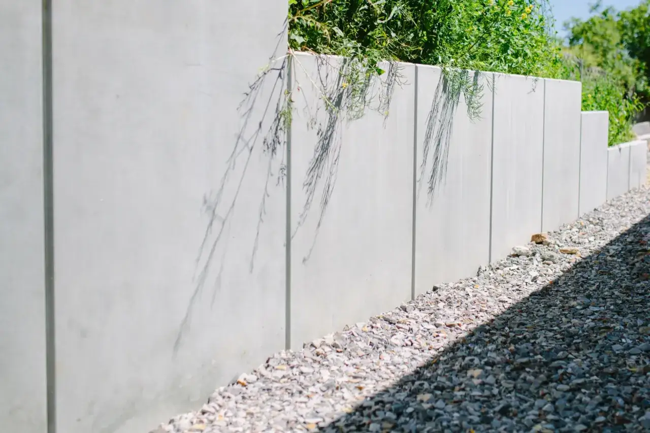 Nowoczesny mur oporowy z betonu, z żwirem u podstawy. Widać cień gałęzi na jego powierzchni. Odległość od granicy działki jest zachowana.