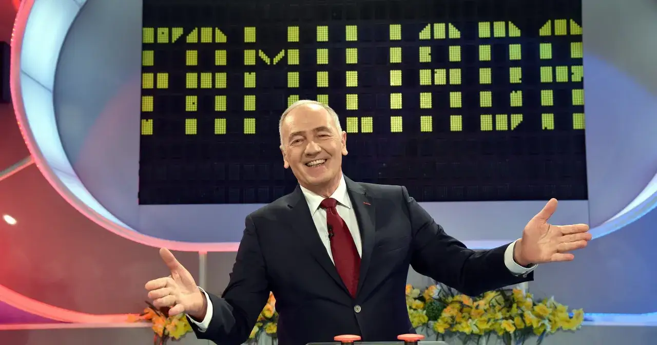Uśmiechnięty prowadzący w garniturze, na tle planszy z zielonymi polami. Chciałby być zdrowy jak Familiada, pełen energii i radości.