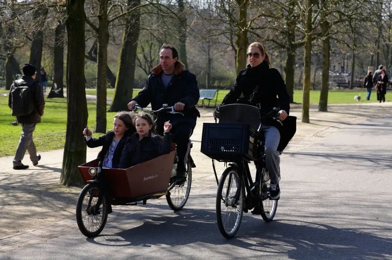Rodzina na rowerach w parku, typowe życie w Holandii. Dzieci w bakfiets, rodzice jadą obok.