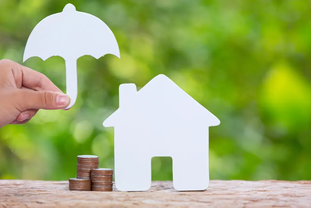 Mano tiene ombrello bianco sopra casa bianca e pile di monete. Rappresenta la sicurezza finanziaria, come fideiussione e deposito cauzionale insieme.