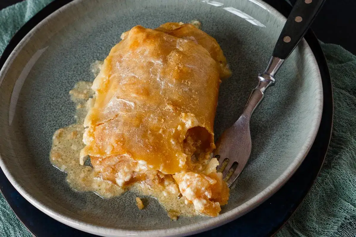 Ein goldbrauner Apfelstrudel mit Eiermilch, der auf einer grauen Platte serviert wird. Eine Gabel steckt im Strudel.