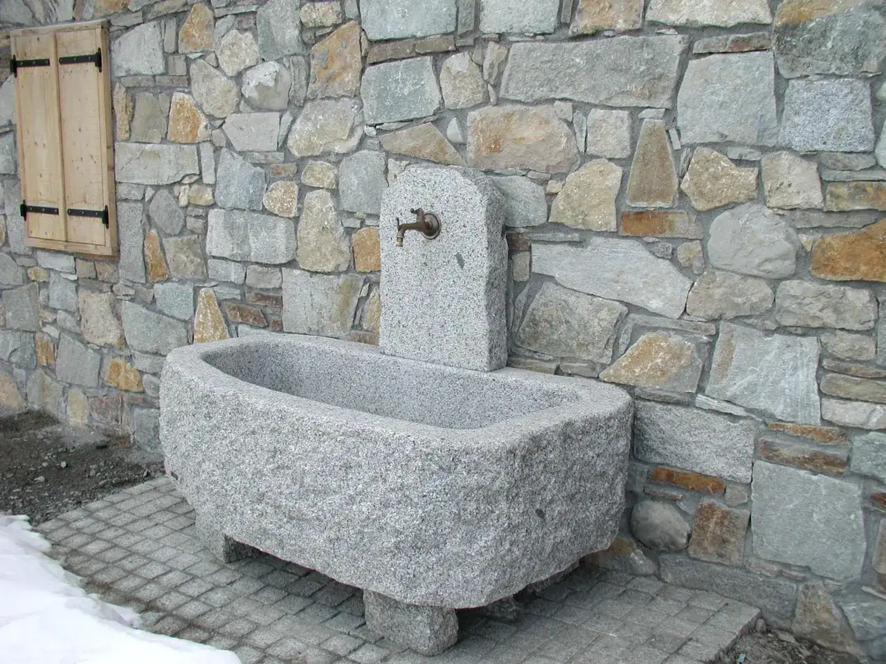 Fontaine en pierre sculpt&eacute;e, avec une bordure de bassin en pierre rustique, adoss&eacute;e &agrave; un mur de pierres s&egrave;ches.
