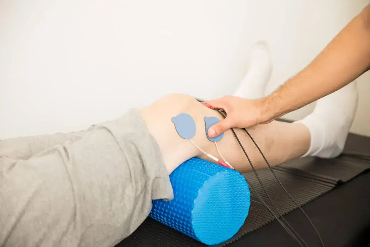 Fizjoterapeuta stosuje elektrody do kolana pacjenta, co może być częścią rehabilitacji po urazie, np. szyciu łąkotki.