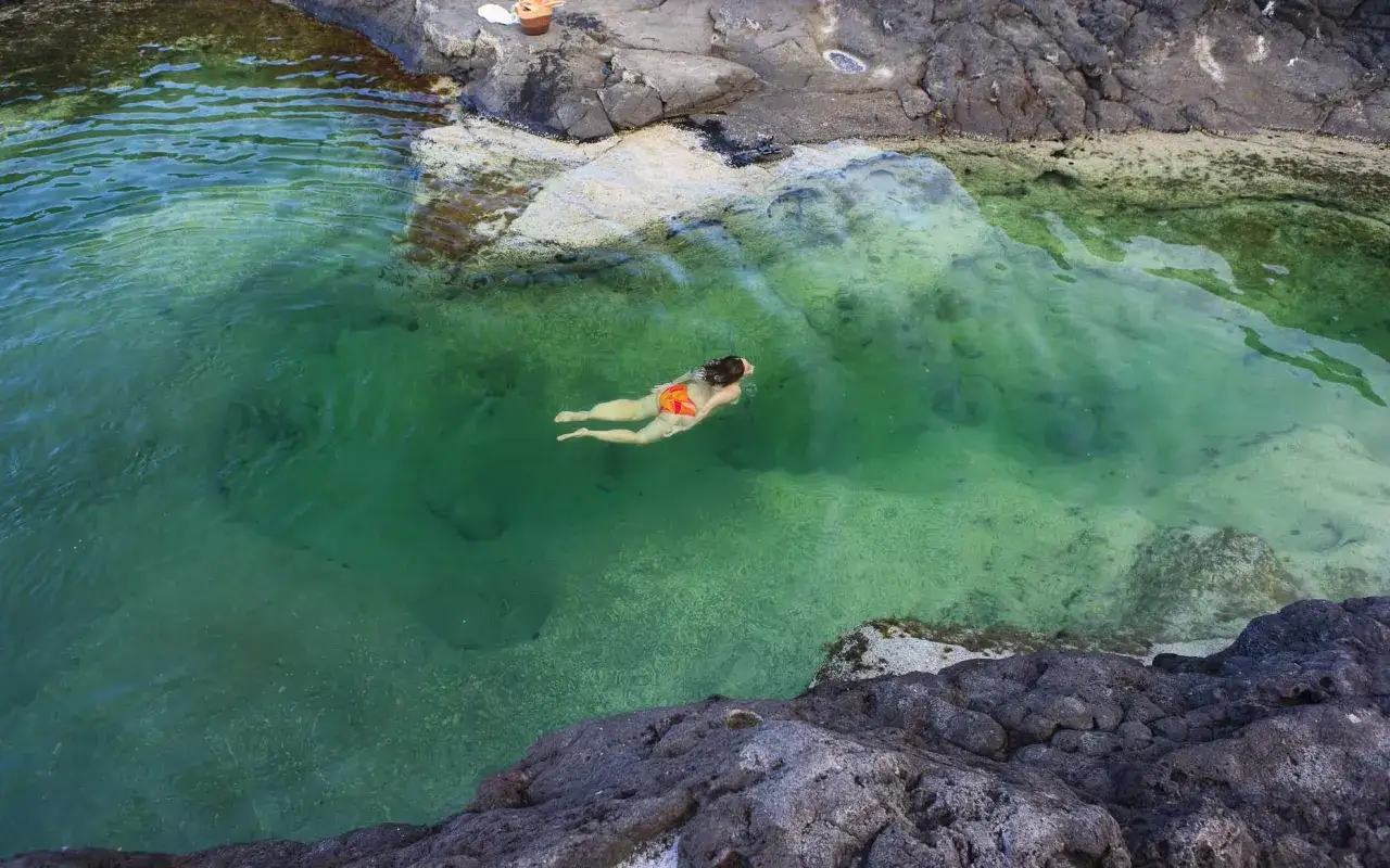 Una mujer nada en una piscina natural de aguas turquesas, rodeada de rocas volcánicas. Un paraíso escondido, como los cocoteros de Lanzarote.