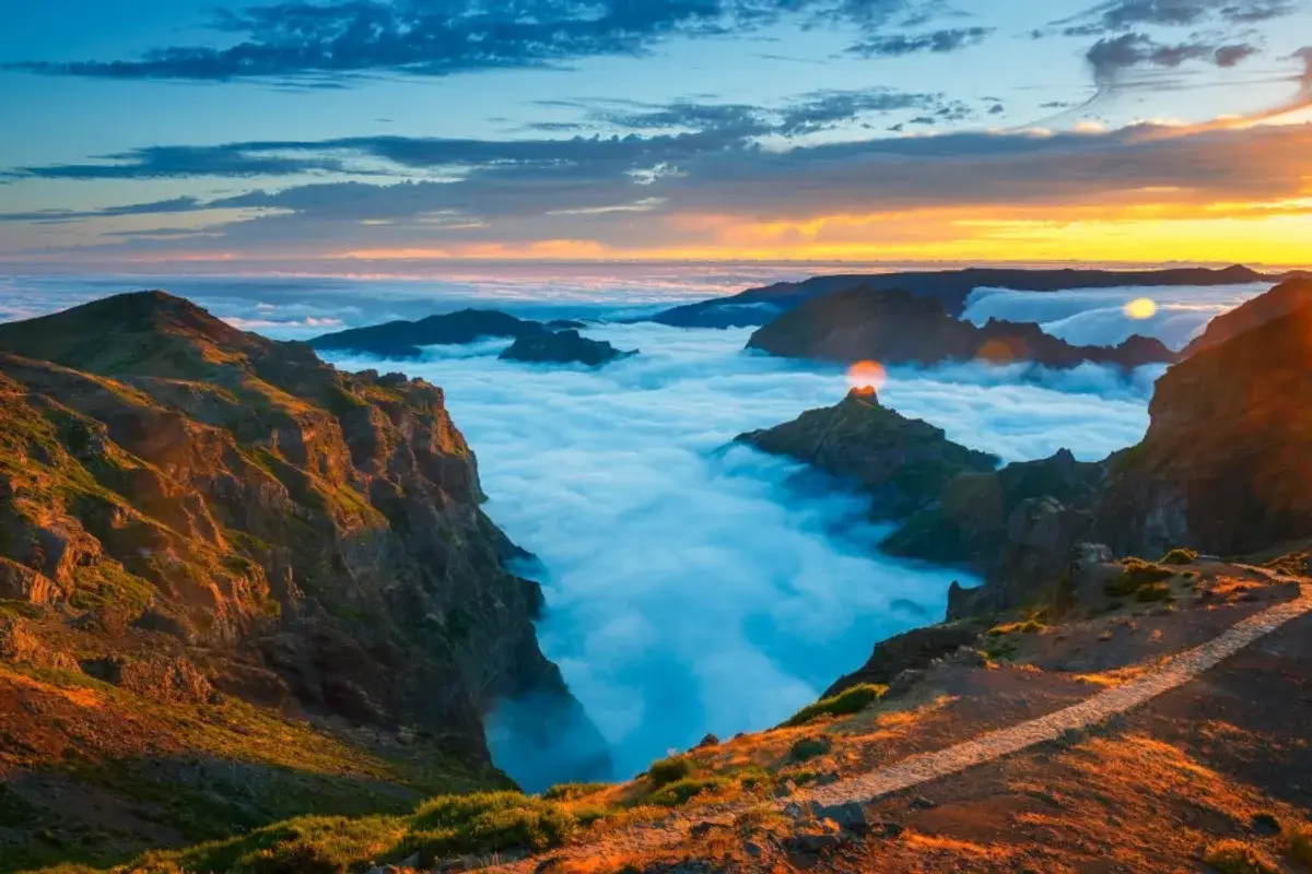 Sonnenaufgang &uuml;ber Wolkenmeer auf Madeira. Bergspitzen ragen wie Inseln aus dem Nebel. Ein Pfad f&uuml;hrt durch die dramatische Landschaft.