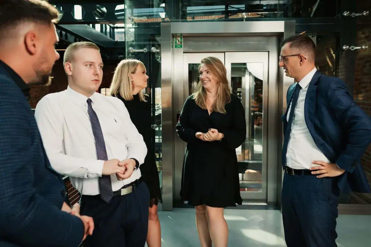 Grupa profesjonalistów w eleganckich strojach rozmawia przed windą w nowoczesnym biurze.